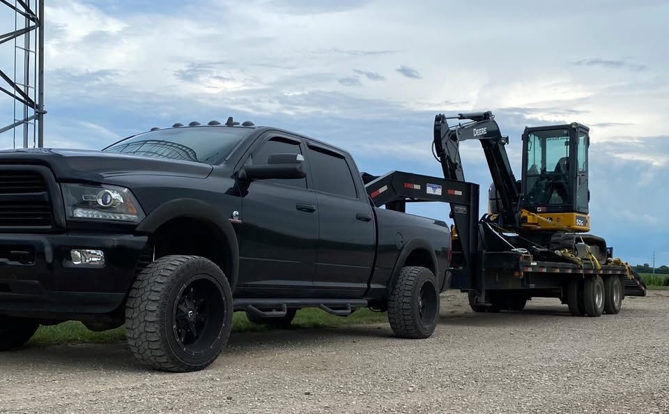 Black pickup truck towing a trailer with a small excavator on a gravel road, overcast sky.