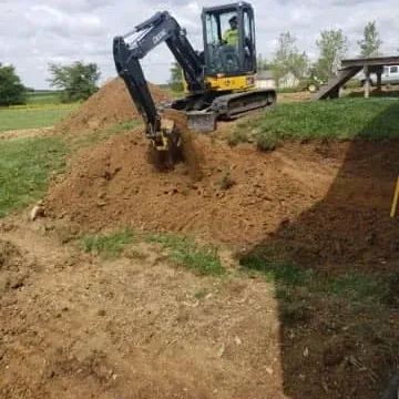 Excavator digging into a dirt pile on a grassy area, operator visible in the cab.