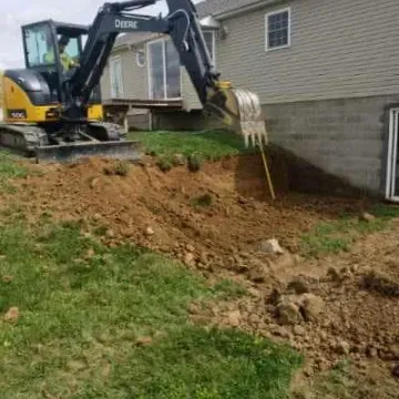 Excavator digging near a house; dirt and grass surround the construction site.