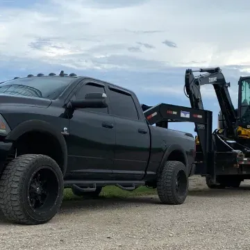 Black pickup truck towing a trailer with a piece of heavy equipment. Overcast sky.