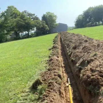 Trench dug in a grassy yard, dirt piled along edges; yellow shovel visible.
