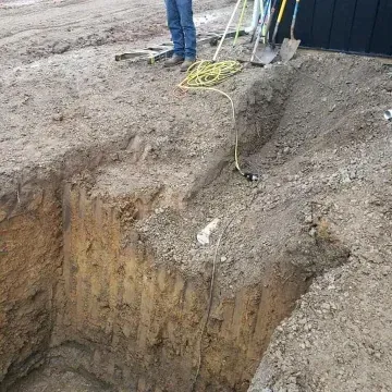A deep excavation pit with a person standing near it, construction tools are scattered.
