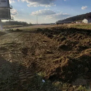 Dirt field with tractor tracks, clouds overhead, commercial buildings and a communications tower in the distance.
