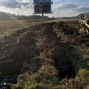 A trench dug in a field with a billboard and construction equipment in the background.