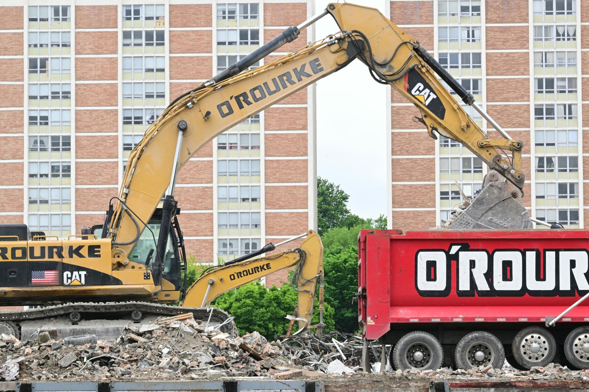 Yellow excavator loading debris into a red O'Rourke dump truck during building demolition.