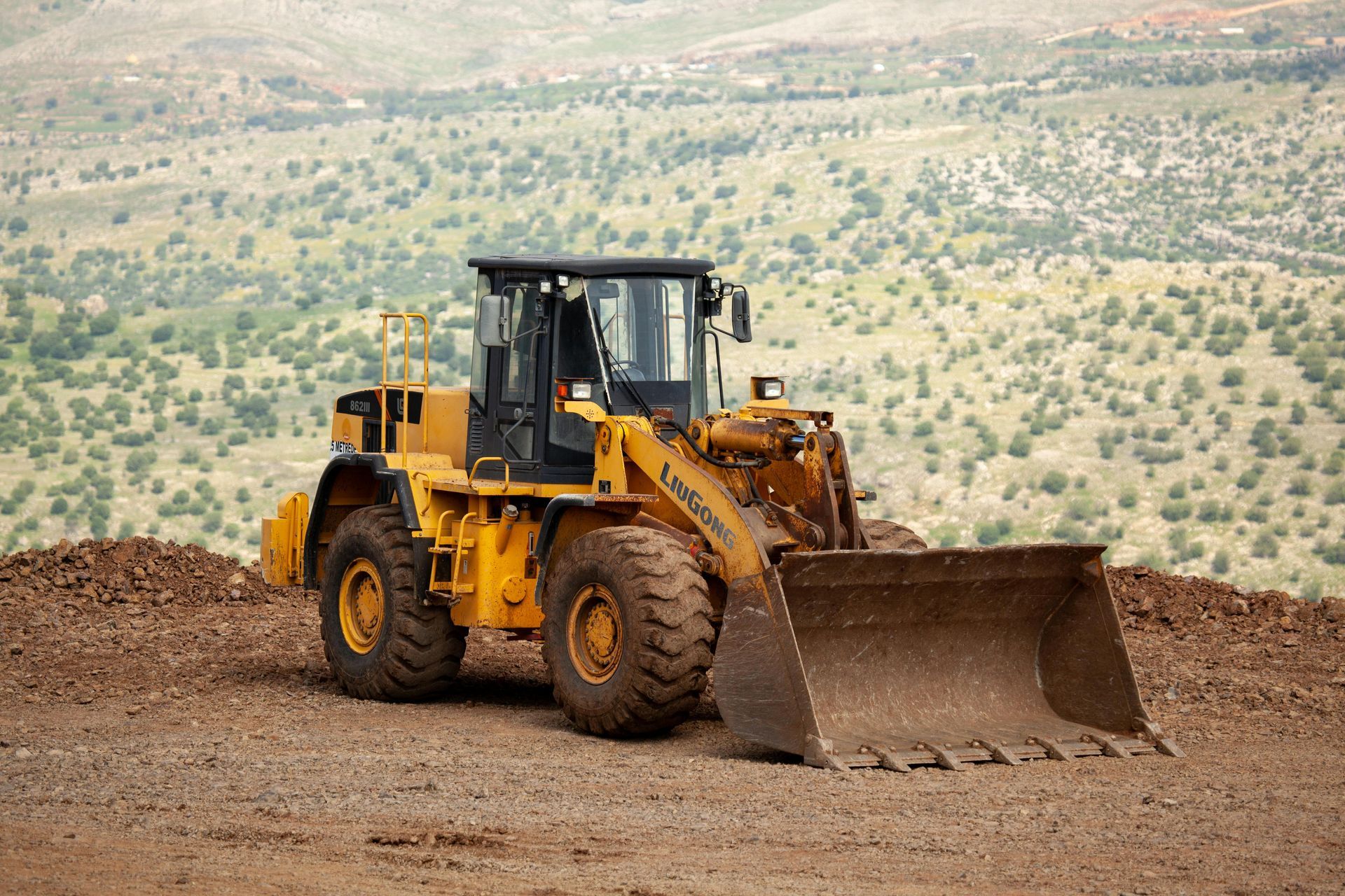 Yellow front-end loader on dirt, with a hilly, sparsely wooded landscape in the background.