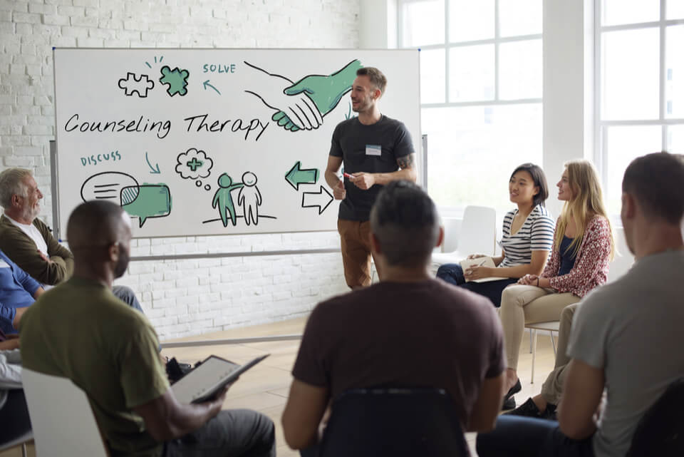 Un gruppo di persone è seduto in cerchio davanti a una lavagna.