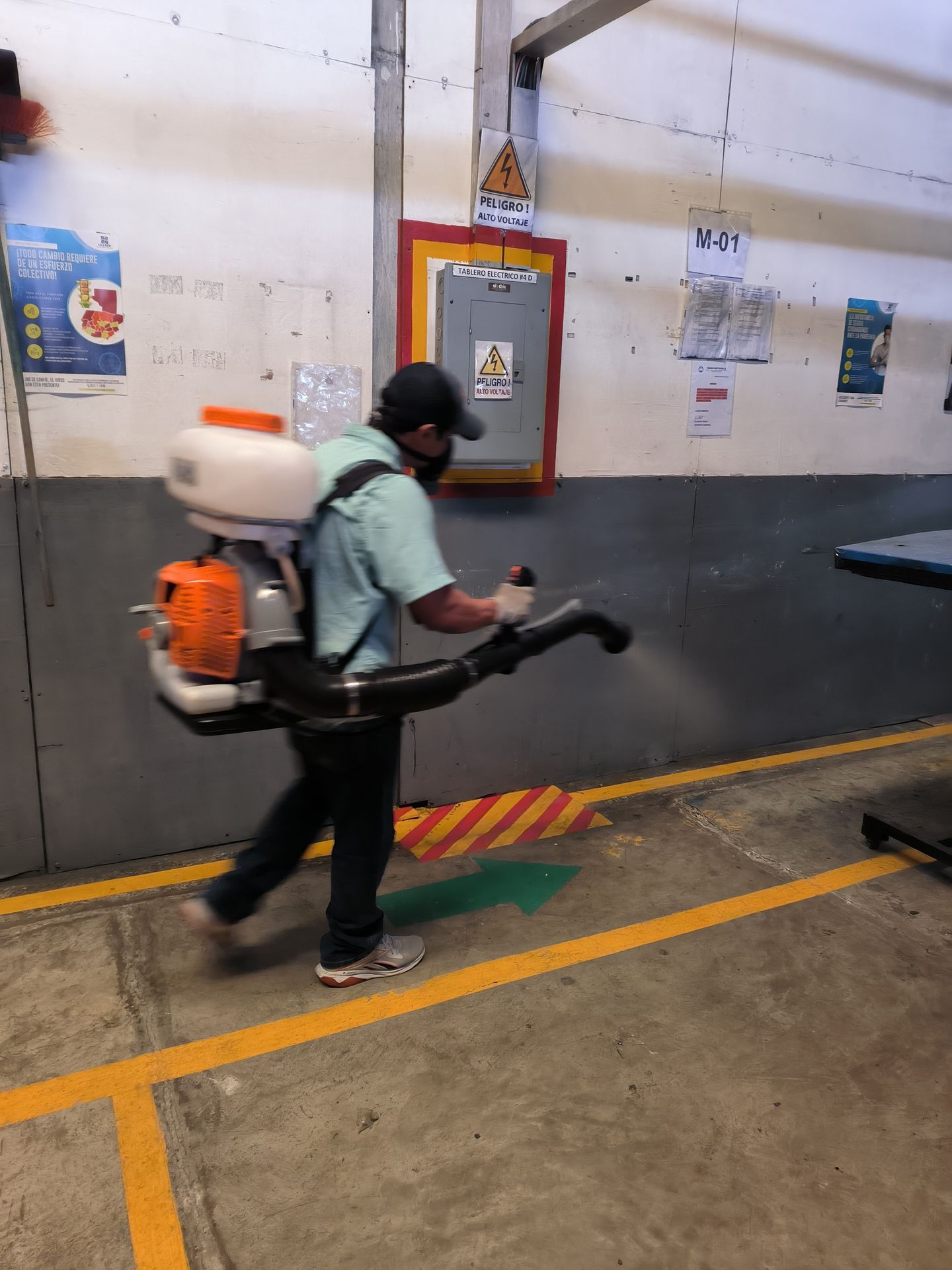 Un trabajador con una camisa azul claro y una mochila pulverizadora desinfecta una pared en una zona industrial.