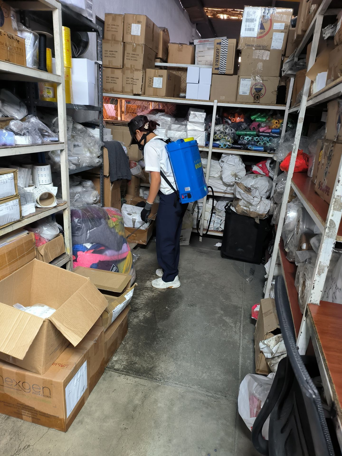 Una persona que lleva una mochila pulverizadora azul desinfecta un almacén lleno de estanterías y cajas de cartón.