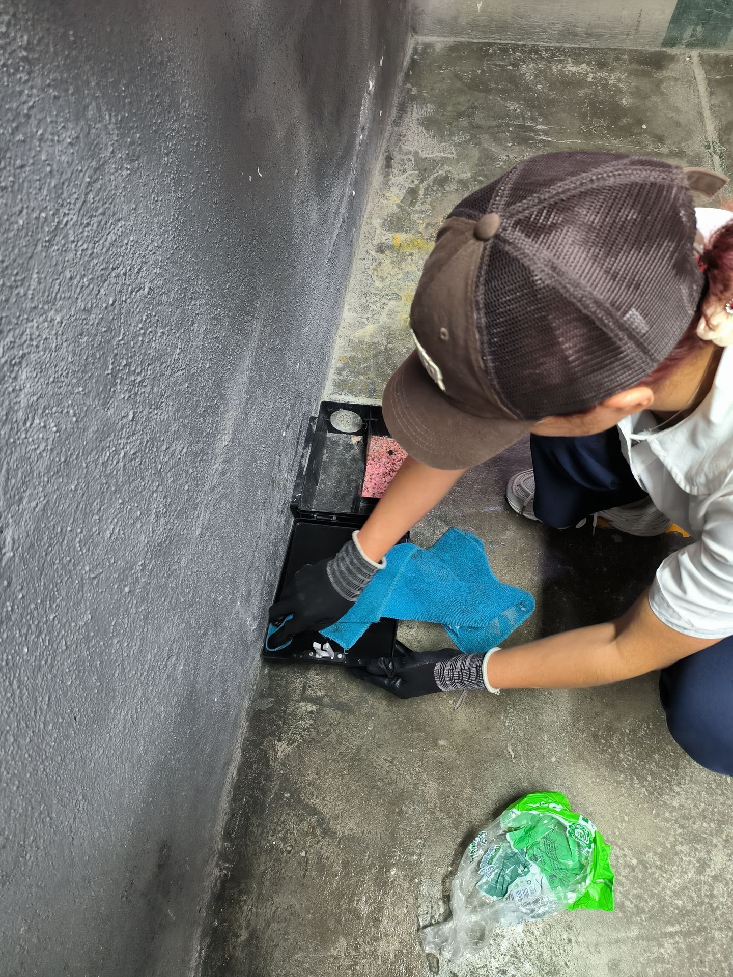 Una persona con gorro y guantes marrones limpia con un trapo azul un desagüe negro cerca de una pared gris.