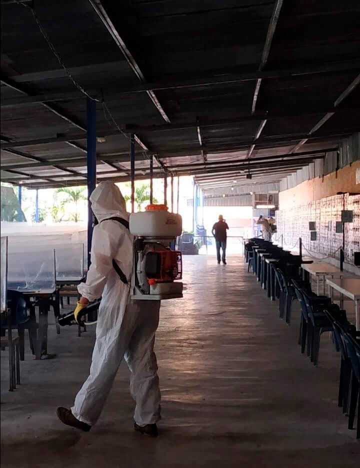 Un trabajador con equipo de protección completo rocía desinfectante en un área al aire libre con filas de sillas y mesas.