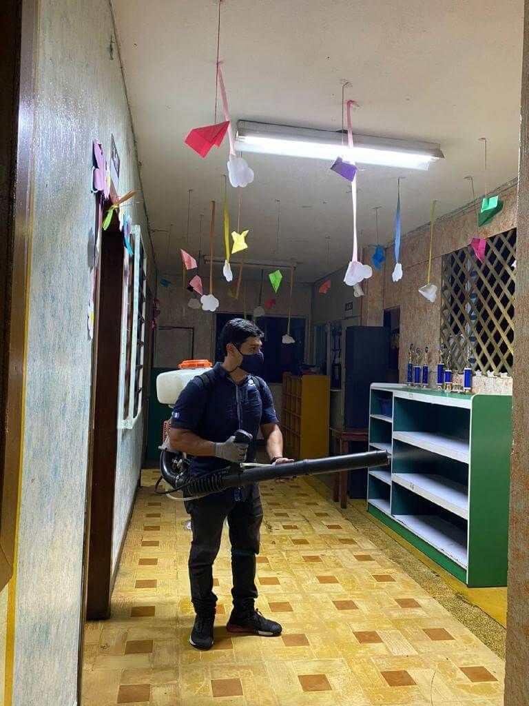 Una persona con mascarilla y ropa oscura utiliza un pulverizador de mochila para desinfectar un pasillo 