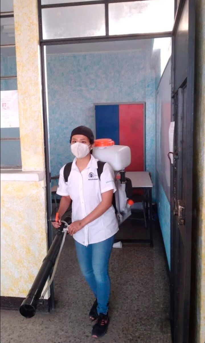 Una persona con camisa blanca, mascarilla y mochila pulverizadora desinfecta la entrada de una habitación.