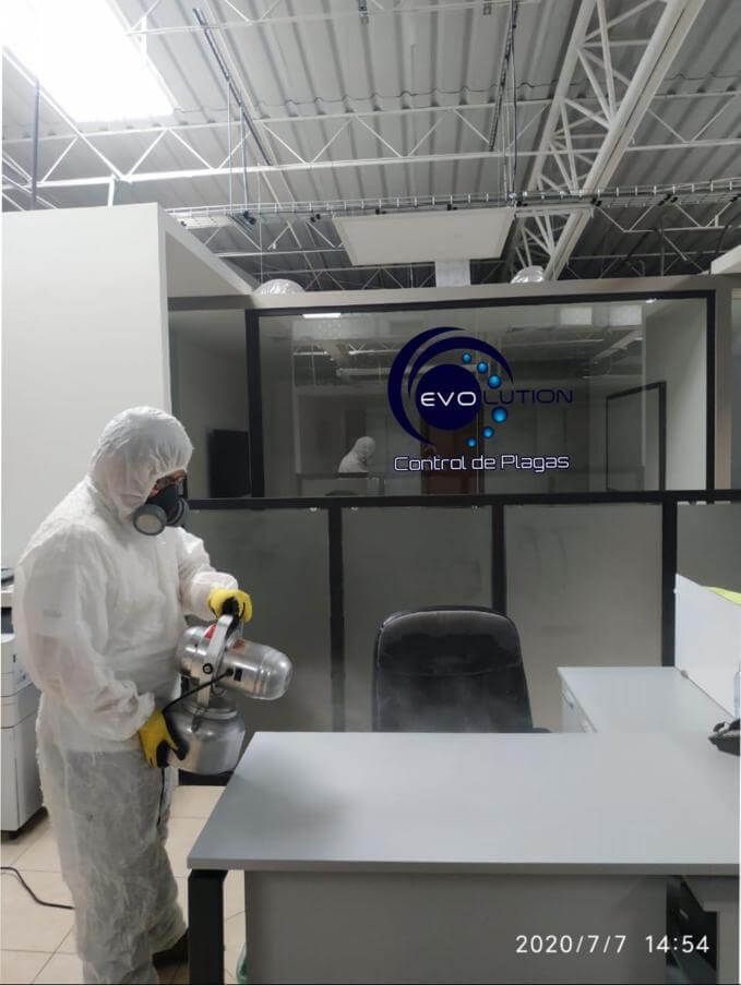 Una persona con equipo de protección completo desinfecta un espacio de oficina rociando una niebla sobre un escritorio.