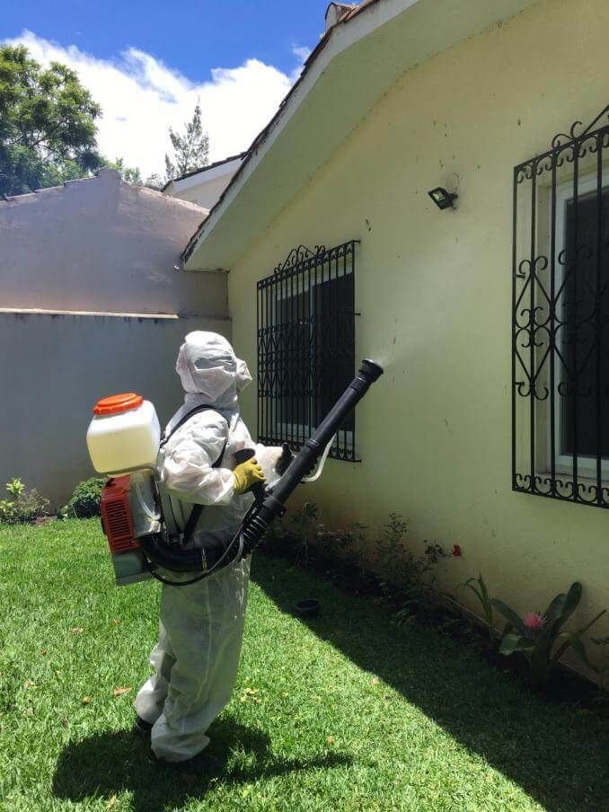 Una persona con equipo de protección completo rocía una niebla desinfectante sobre la fachada de una casa blanca.
