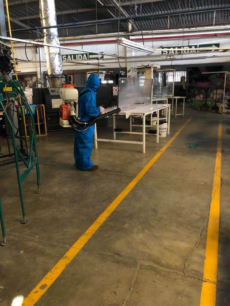 Una persona con un traje protector azul utiliza un pulverizador de mochila para desinfectar un espacio 