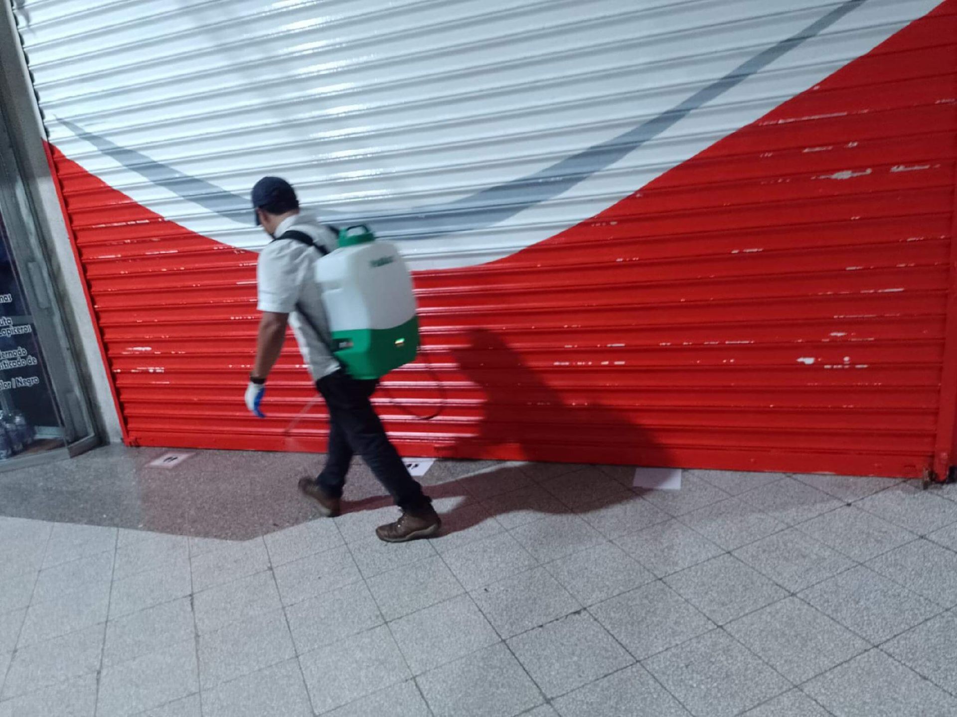 Una persona con uniforme blanco y una mochila pulverizadora camina frente a una puerta enrollable roja y blanca cerrada.