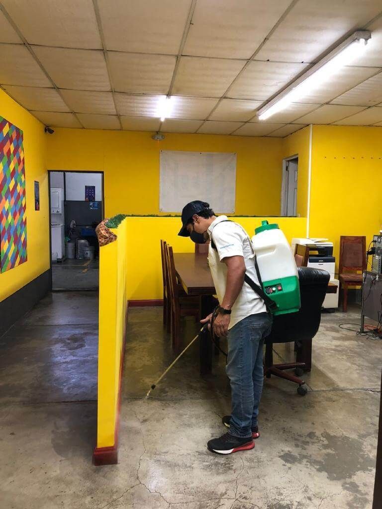 En una oficina de color amarillo, una persona rocía el suelo con un tanque desinfectante portátil.