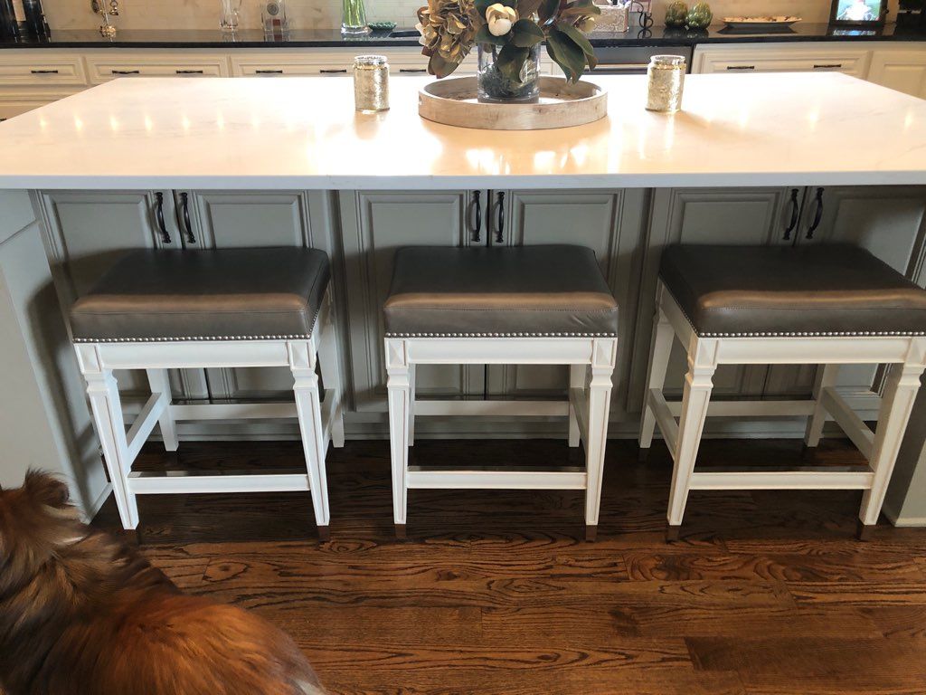 A dog is standing in front of a kitchen island with three stools.