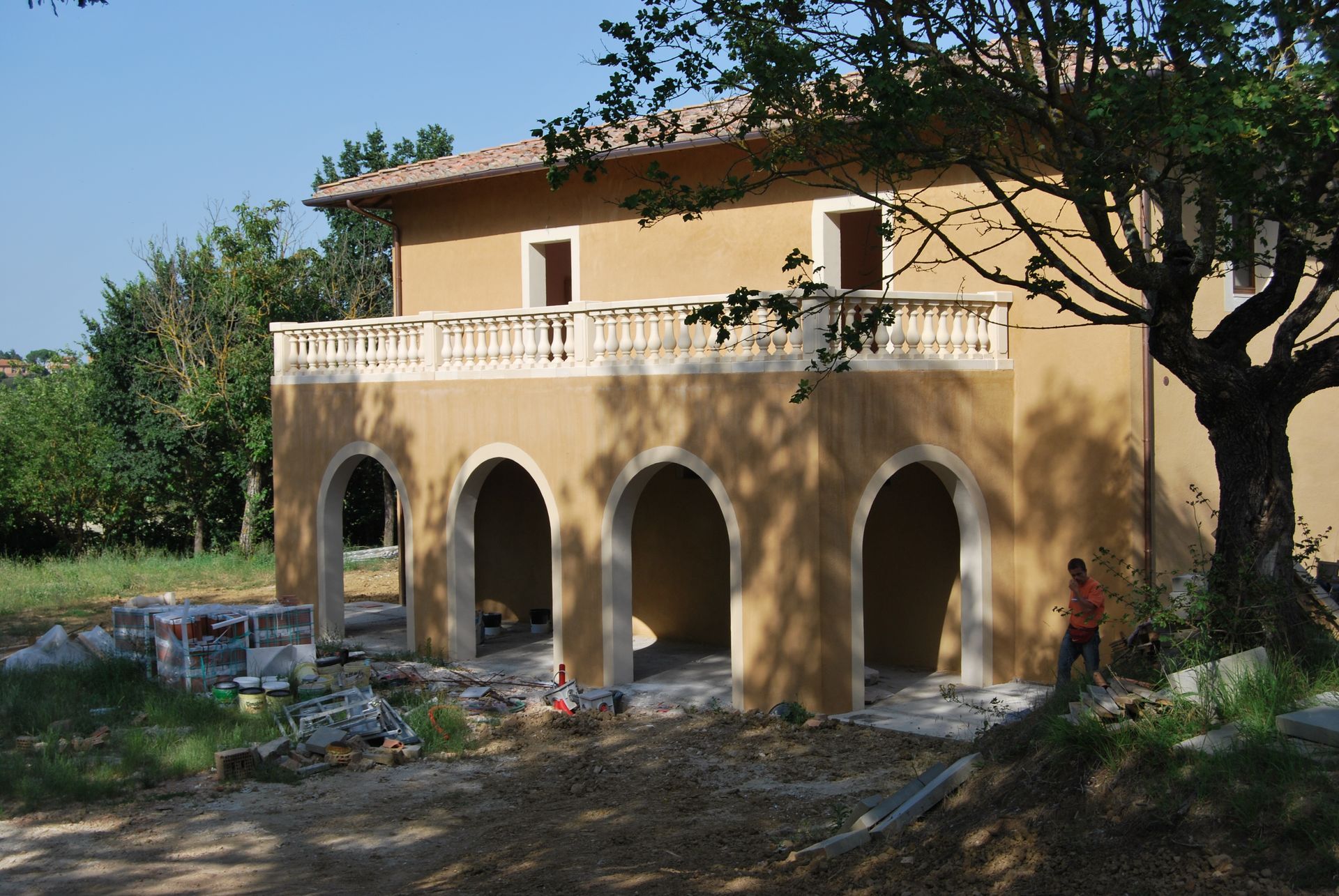 Edificio a due piani con archi al piano terra, balcone in alto, uomo in piedi vicino alla costruzione, giornata di sole.