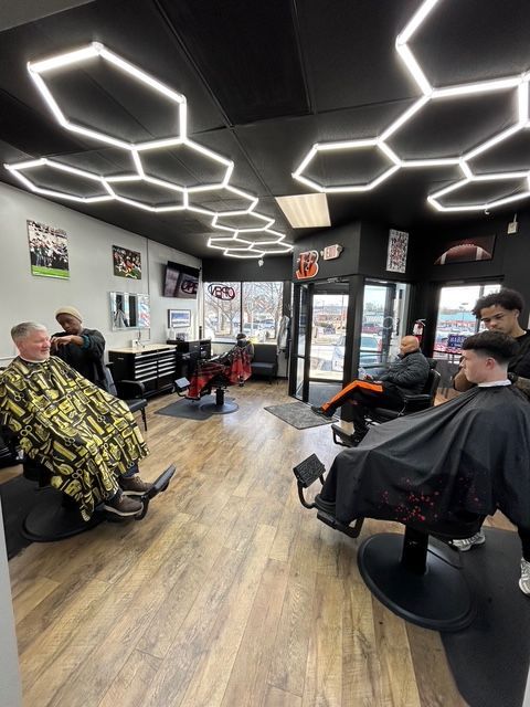 Barbershop interior with customers getting haircuts. Black ceiling with hexagon lights, wood-look floor.