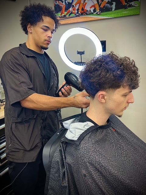 Barber using a hair dryer on a client's curly hair. The client has some purple hair dye. Salon setting.