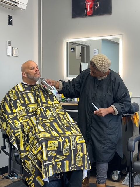Barber trimming a customer's beard in a barbershop. The customer wears a patterned cape.