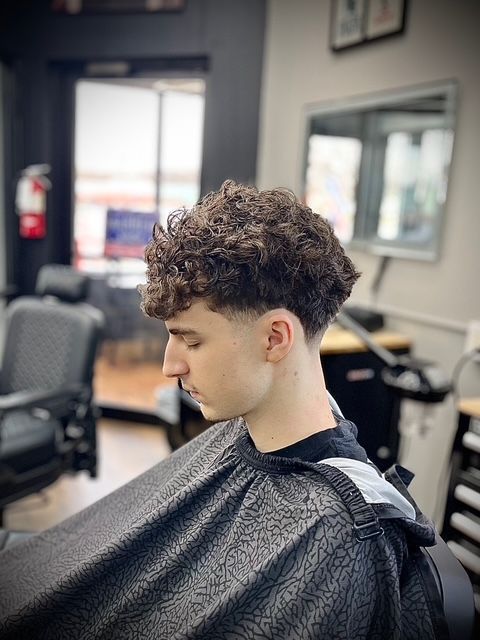 Man with curly hair gets a haircut in a barbershop. He wears a cape and looks down. Faded sides.