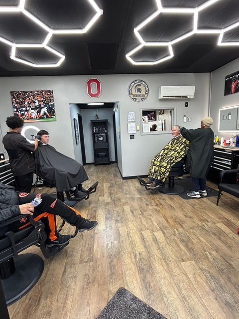 Inside a barbershop with three clients getting haircuts. Overhead, honeycomb-shaped lights illuminate the room.