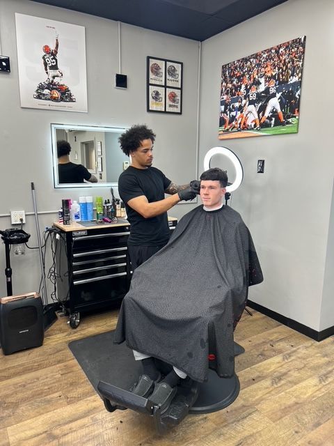 A barber giving a haircut to a client in a barbershop with sports-themed wall art.