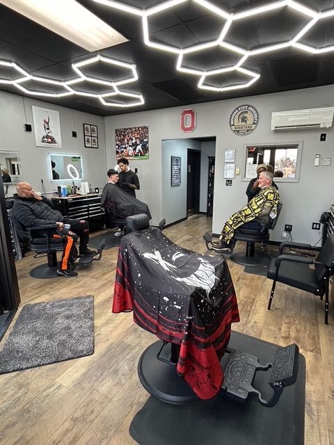 A barbershop interior with several patrons getting haircuts, modern hexagonal ceiling lights, and wooden floors.