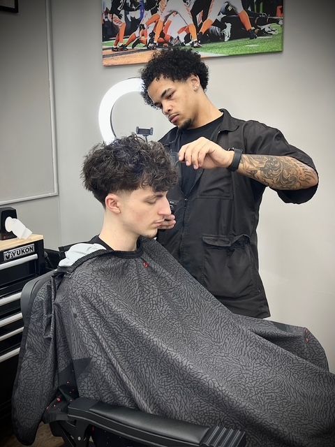 Barber cutting client's hair in a salon. The barber is wearing black and has arm tattoos. The client is seated and wearing a cape.