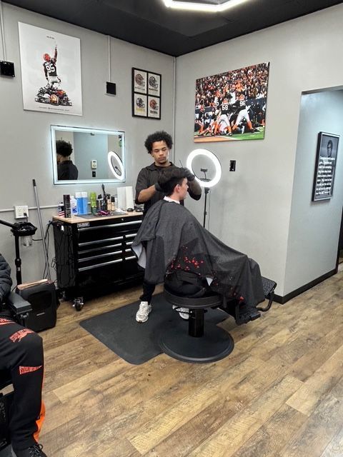 A barber giving a haircut in a modern shop with sports-themed art.