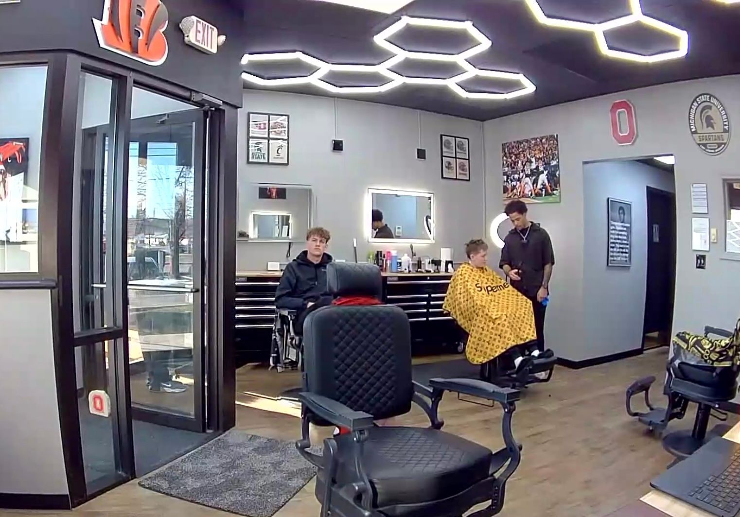 A barbershop interior with two customers getting haircuts. One man is by a barber wearing a yellow cape.