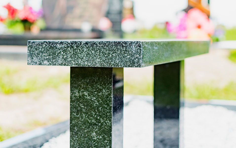 Black granite bench in a cemetery, close-up shot, with grass and flowers in the background.