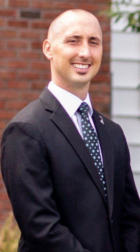 Man in a black suit and tie smiles, standing in front of a brick building.