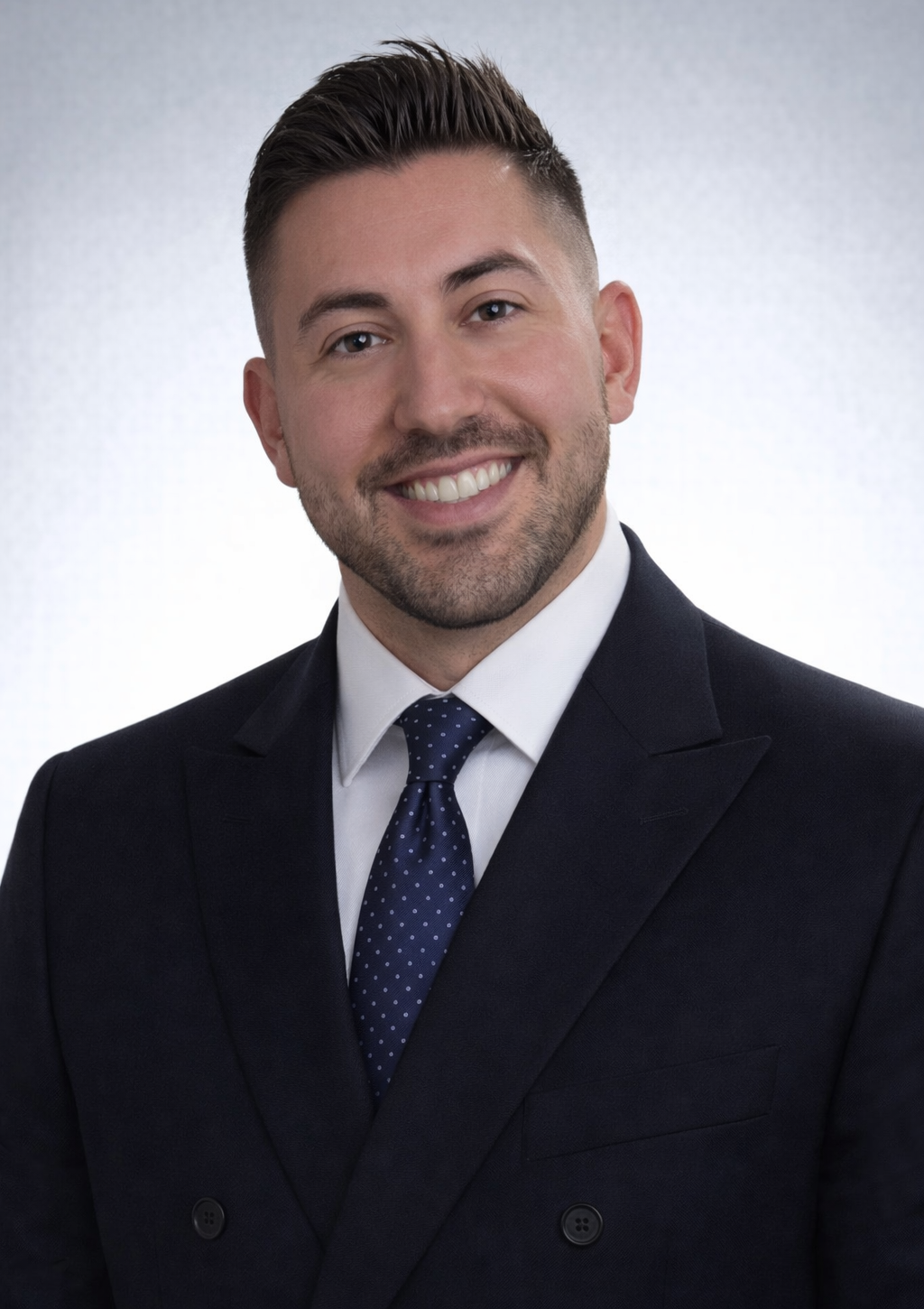 Man in a dark suit with a tie smiles confidently, a studio portrait with a light background.
