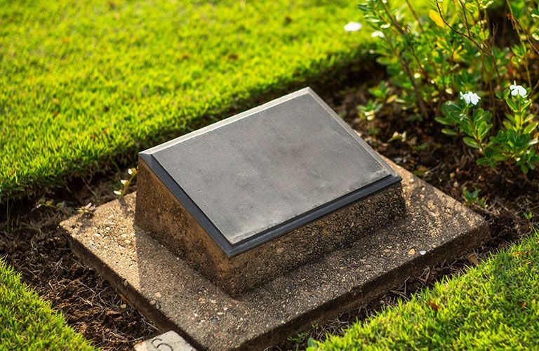 Grave marker: a dark rectangular plaque on a stone base, surrounded by green grass and plants.