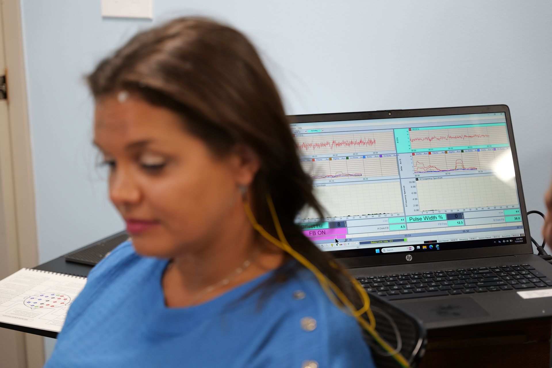A woman with electrodes on her head is seated in front of a laptop displaying data. She is wearing a blue shirt, likely undergoing a medical exam.
