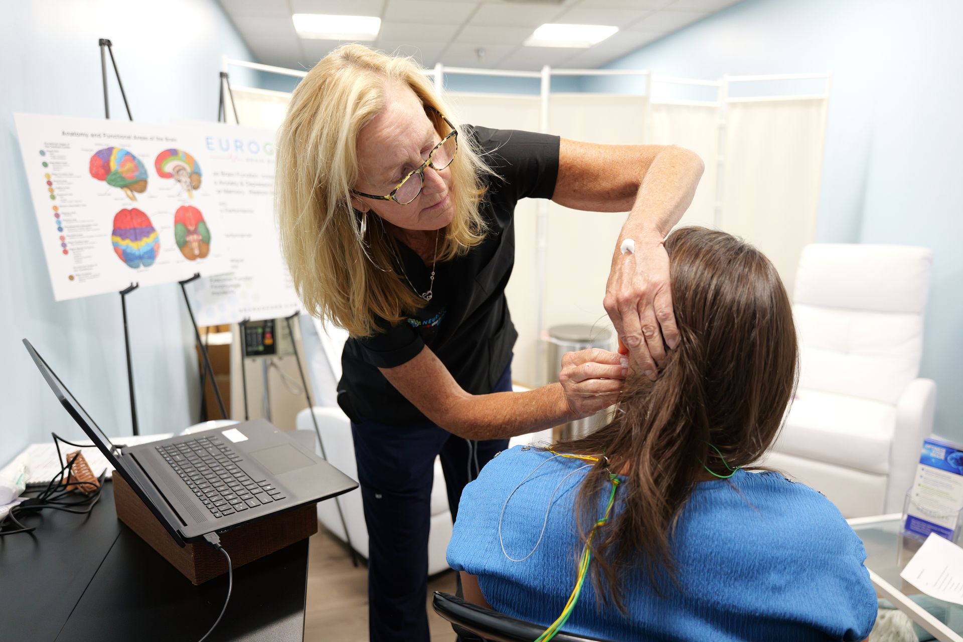 A woman attaches wires to a patient's head, next to a laptop and brain diagram in a medical office. The patient sits in a chair.