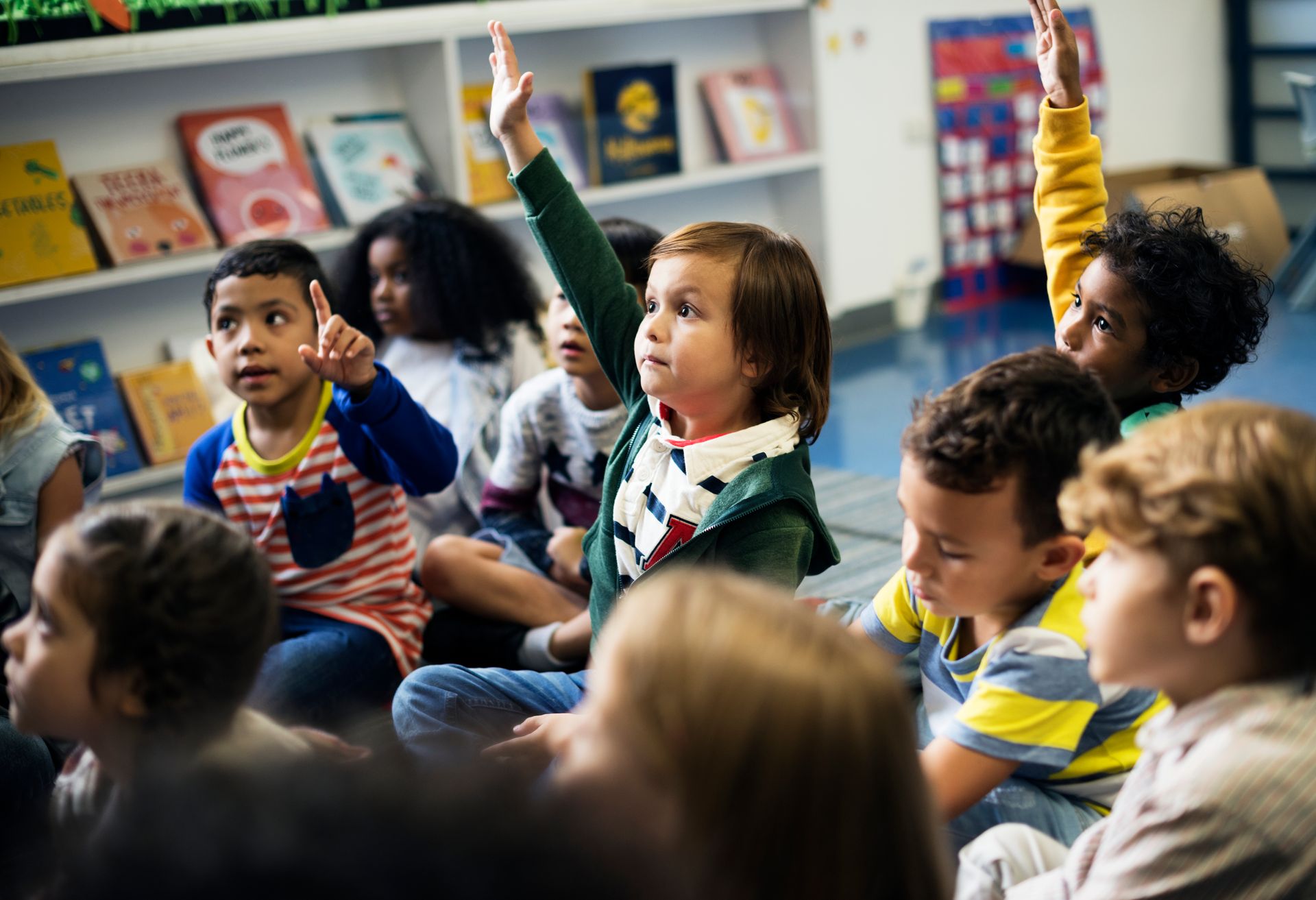 A group of children are sitting on the floor in a classroom raising their hands to answer a question.