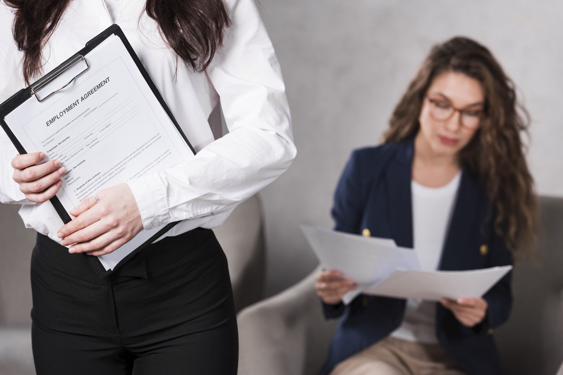 A woman is holding a clipboard and a woman is sitting in a chair holding papers.