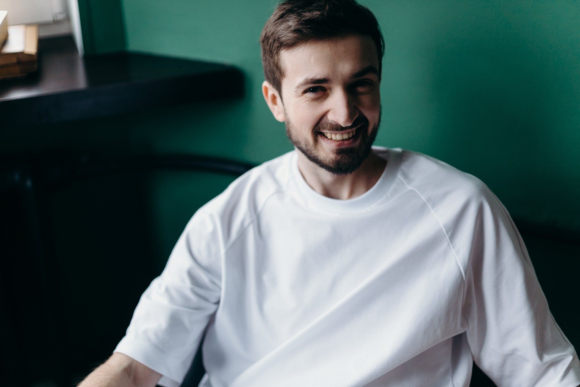 A man with a beard is wearing a white t-shirt and smiling.