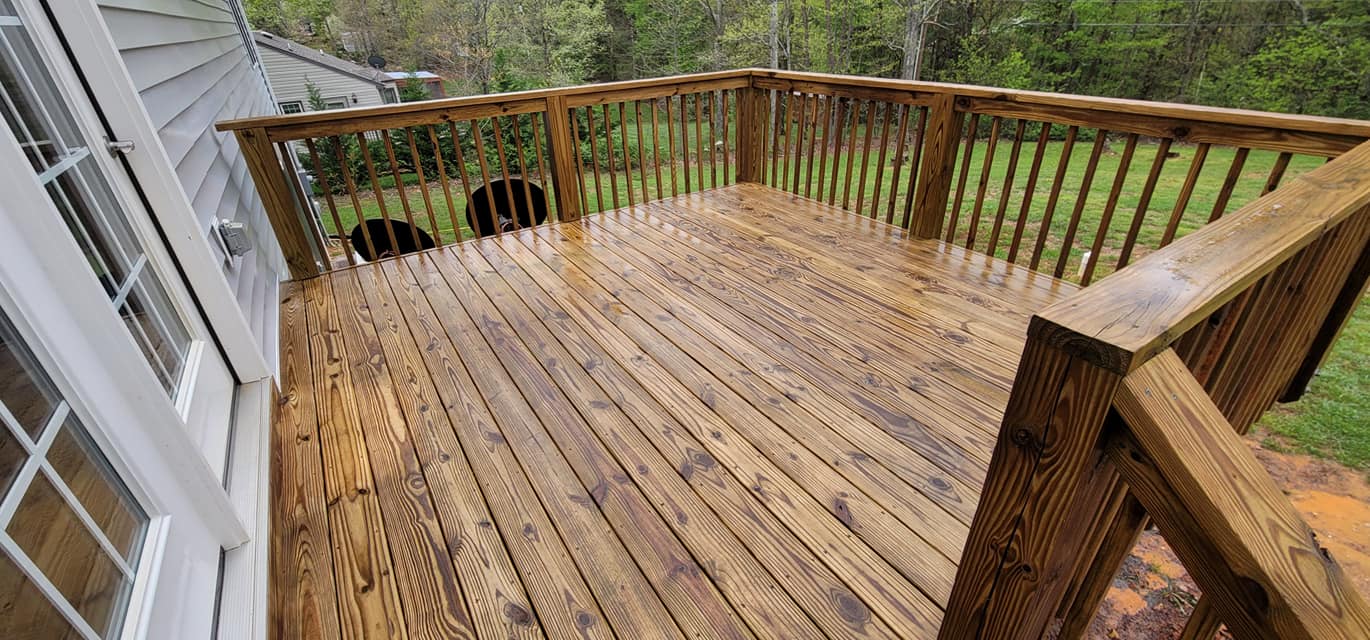 A wet, wooden deck with a railing and two dogs in the background, alongside a white house and greenery.