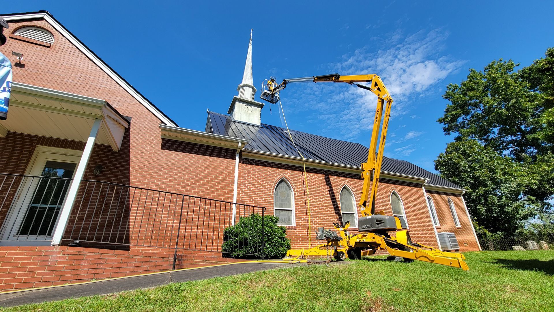A cherry picker spraying a church roof. Yellow machine, red brick building, blue sky.