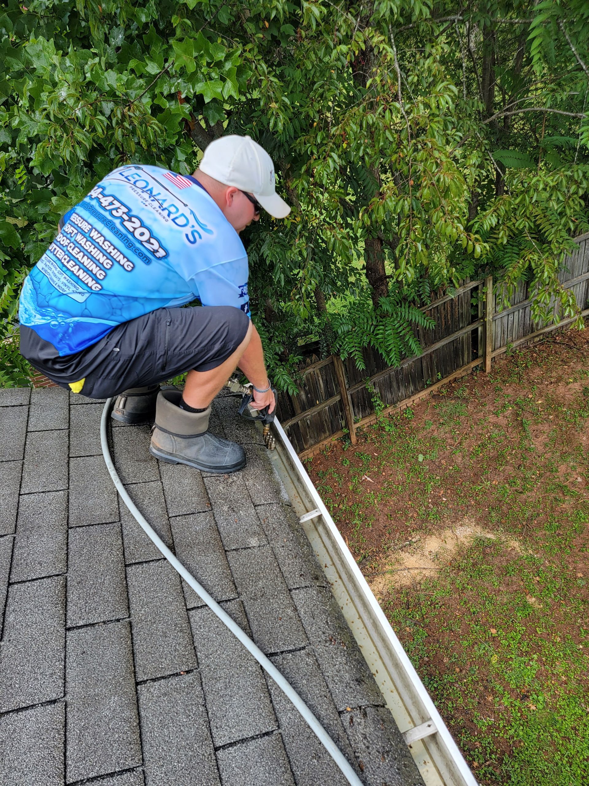 Man cleaning gutters on a roof wearing a light blue shirt, white hat, and black shorts in front of a leafy background.
