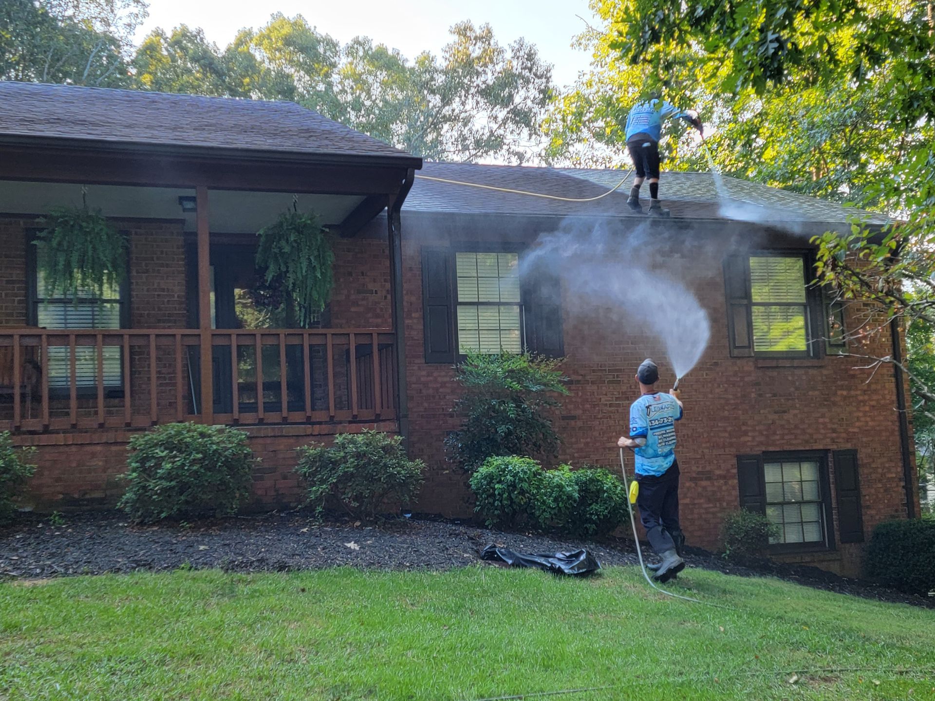 Two people power washing a brick house roof, spraying water, sunny outdoor setting.
