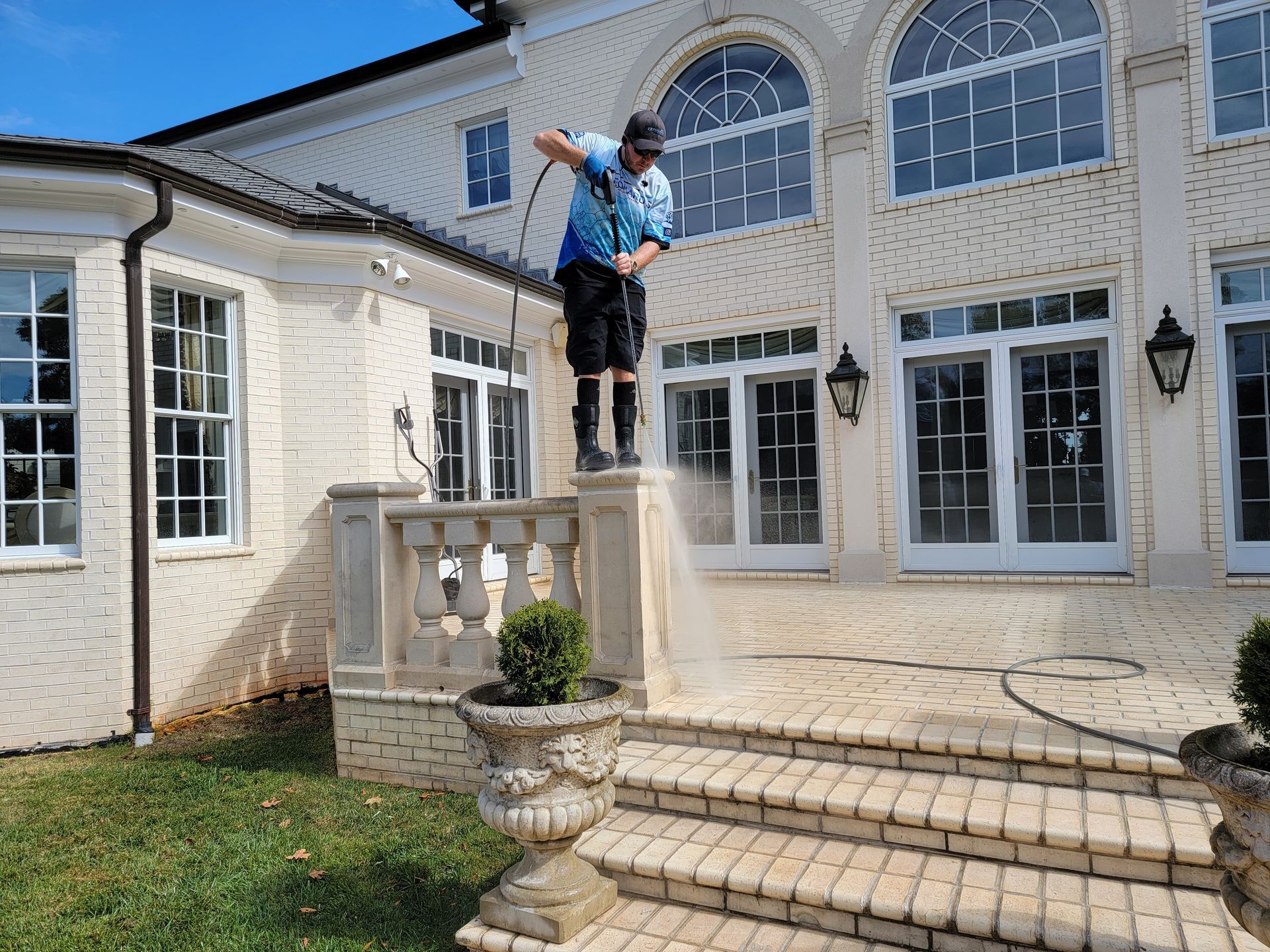 Man pressure washing a white brick house's patio, wearing shorts, on a sunny day.