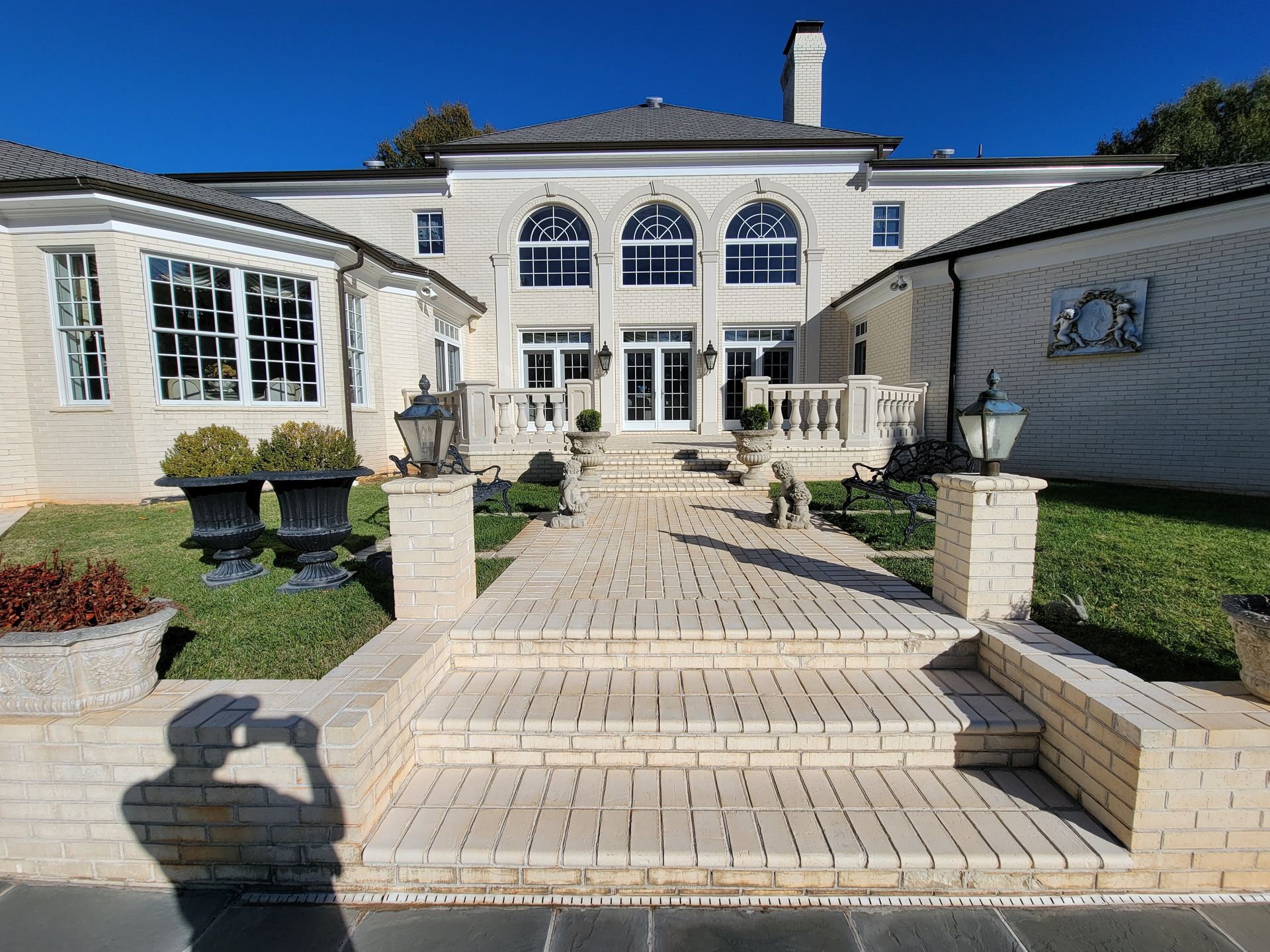 Elegant light-colored mansion with arched windows, brick pathway, and manicured lawn, under a clear sky.