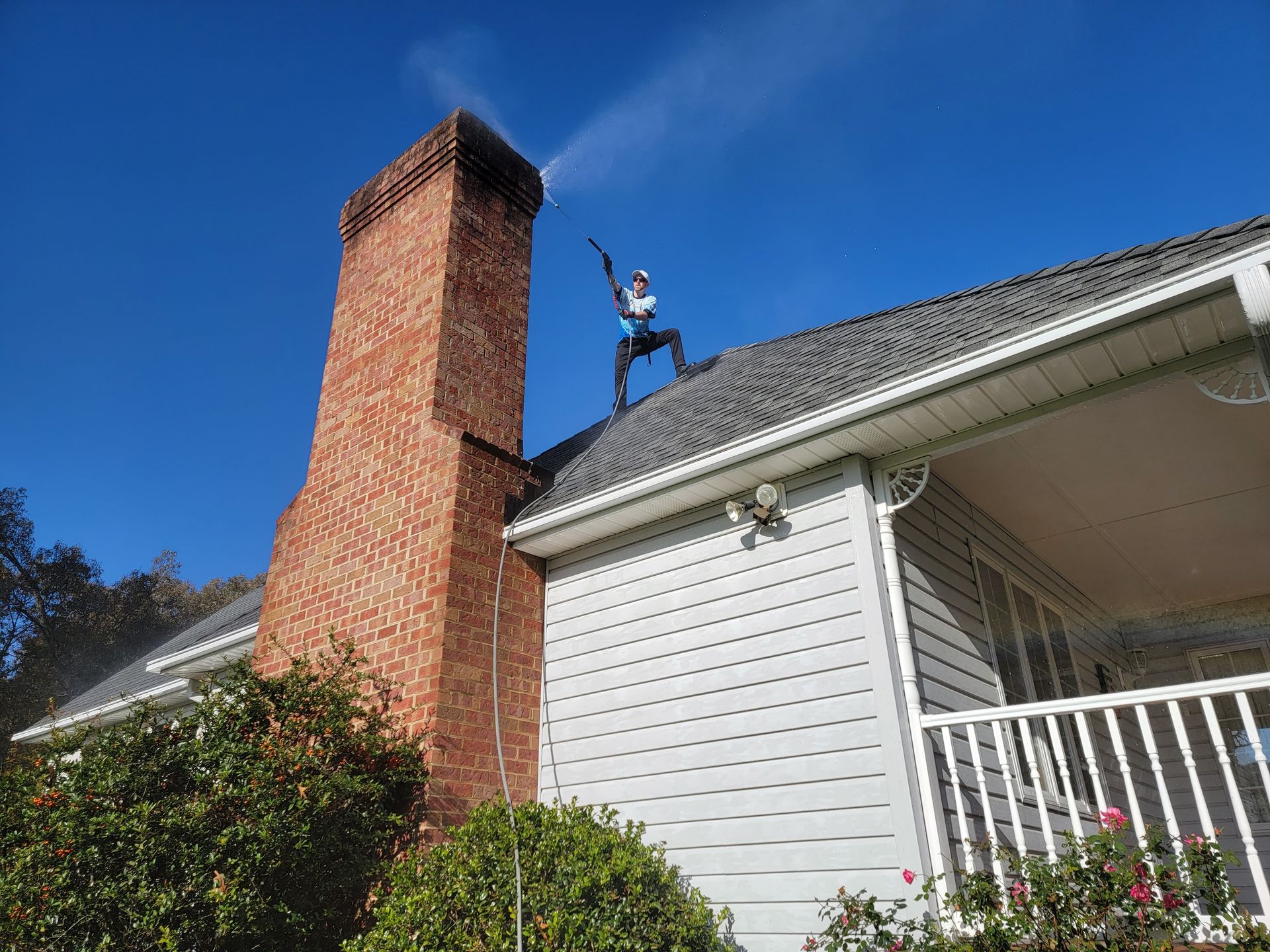 Person on a roof spraying water from a chimney. Brick chimney, blue sky, and a white house.